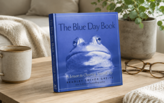 The Blue Day Book by Bradley Trevor Greive on a wooden table, blue cover with a close-up frog photograph, subtitle "A Lesson in Cheering Yourself Up," styled in a cosy home setting.