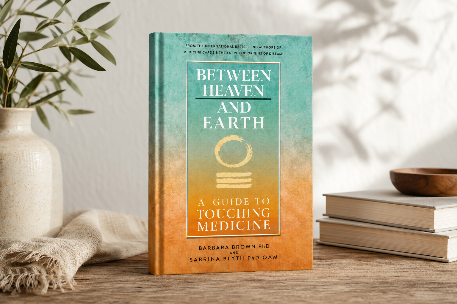 Between Heaven and Earth: A Guide to Touching Medicine by Barbara Brown PhD and Sabrina Blyth PhD OAM. Book cover with turquoise-to-orange gradient displayed on a wooden surface with a cream ceramic pot, green foliage, and stacked books, styled with natural light and coastal minimalist décor.